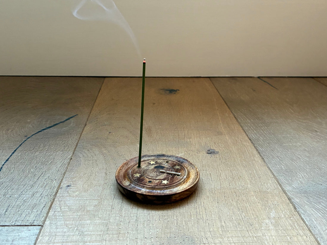 A single EcoIncense stick burning gently in a wooden holder on a natural oak floor, with soft smoke rising — symbolising calm and mindfulness.